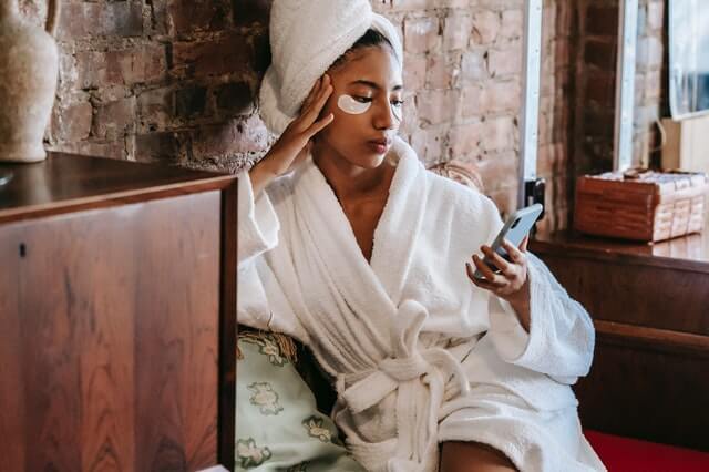 A woman is using Beauty booking system at her mobile phone with bathrobe and face mask on her.